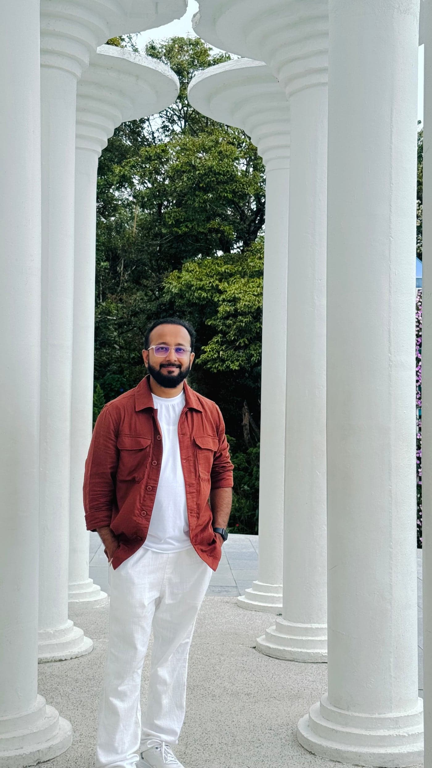 Akshay Katkade standing between tall white columns, wearing a rust overshirt and white trousers.
